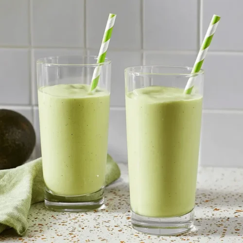 Two glasses of fresh avocado smoothie on a kitchen counter with whole and halved avocados.