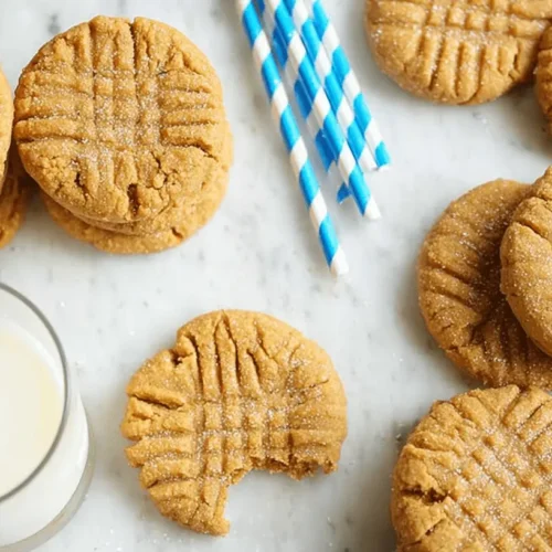 Overhead view of golden-brown Classic Peanut Butter Cookies with a distinctive criss-cross pattern, some with a bite taken out, served alongside a glass of milk and striped straws on a marble surface.