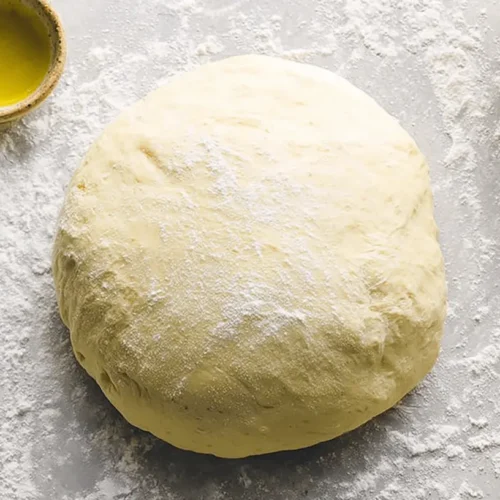 Ball of pale yellow dough dusted with flour, surrounded by olive oil and salt, ready for an easy pizza dough recipe.