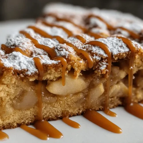 A delicious slice of apple cake generously topped with powdered sugar and caramel drizzle on a white plate.
