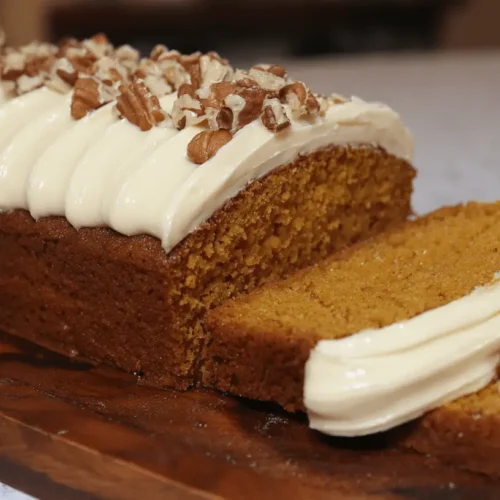 Delicious pumpkin cake loaf, generously frosted with cream cheese and topped with pecans, presented on a dark wooden board.