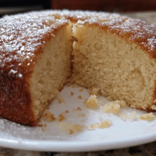 A delicious golden-brown gluten free cake dusted with powdered sugar, with a slice removed revealing its moist, pale yellow interior on a white plate.