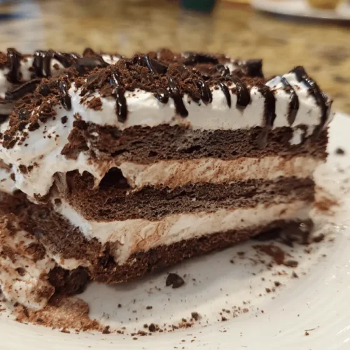 Decadent layered chocolate icebox cake slice with whipped cream and chocolate shavings on a white plate.